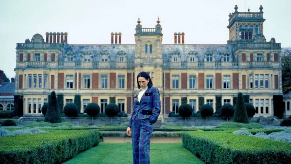 A person in a plaid suit stands in a manicured garden with a large, ornate mansion in the background.