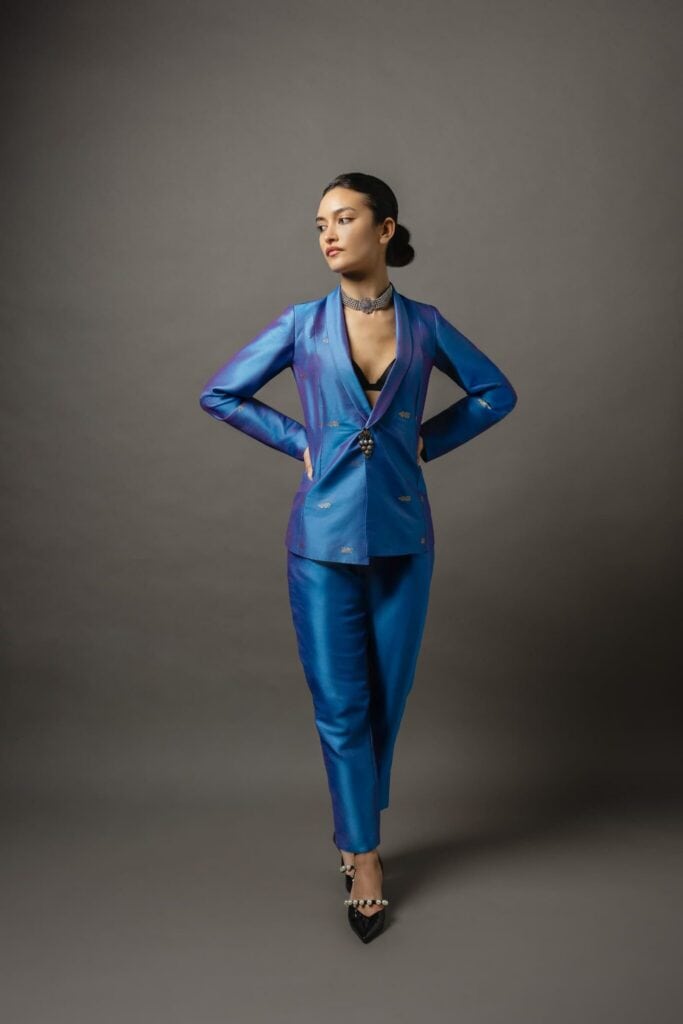A woman stands against a gray background wearing a shiny blue pantsuit with a deep neckline, black heels, and a necklace, with her hands behind her back.