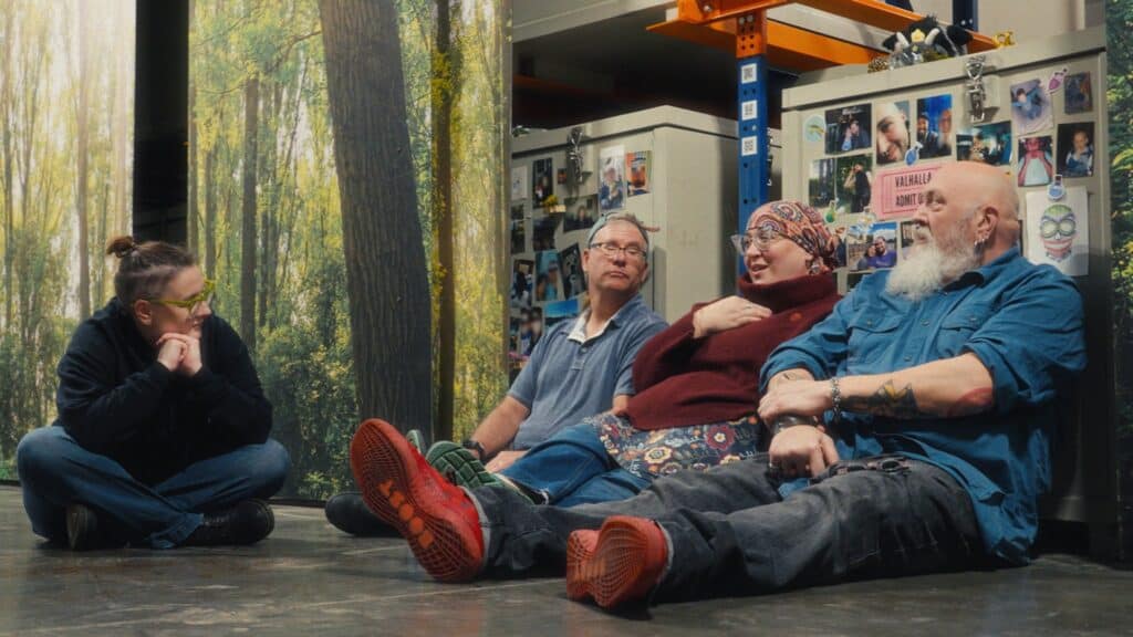 Four people sit on a floor in front of storage cabinets, talking and smiling, with forest-themed panels and photos on the wall behind them.