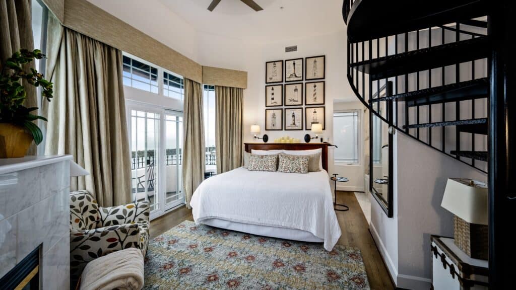 A bright bedroom with a white bed, patterned armchair, spiral staircase, framed art above the headboard, large windows, and a balcony offering views of Bellingham Bay—perfect for a tranquil Lighthouse Stay.