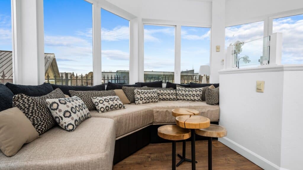 A modern sunroom with large windows overlooking Bellingham Bay, a beige sectional sofa with patterned pillows, and three round wooden nested tables on a hardwood floor—perfect for a Lighthouse Stay experience.