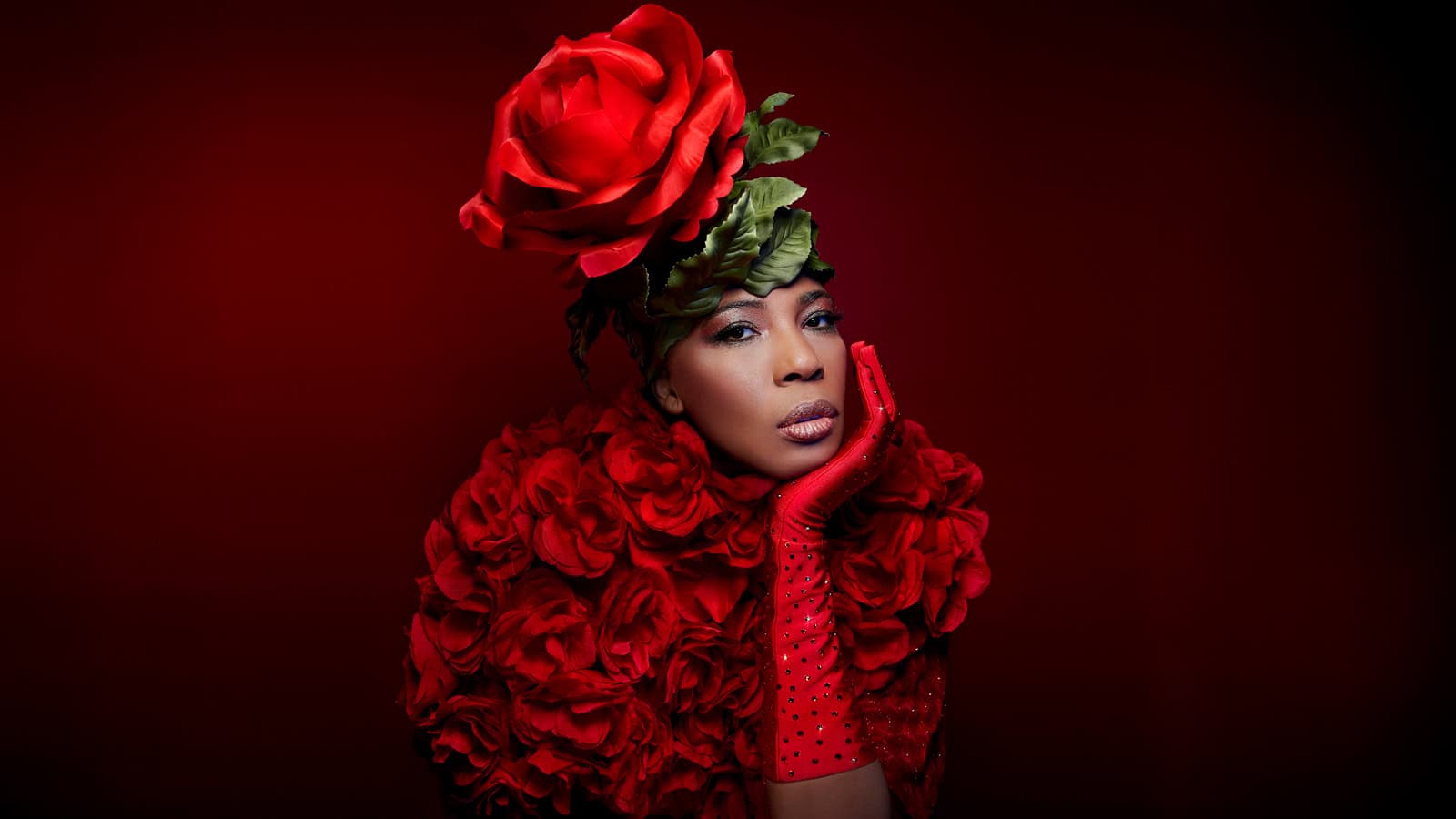 Person wearing a large red rose headpiece, red floral outfit, and red glove, posing against a matching red background.