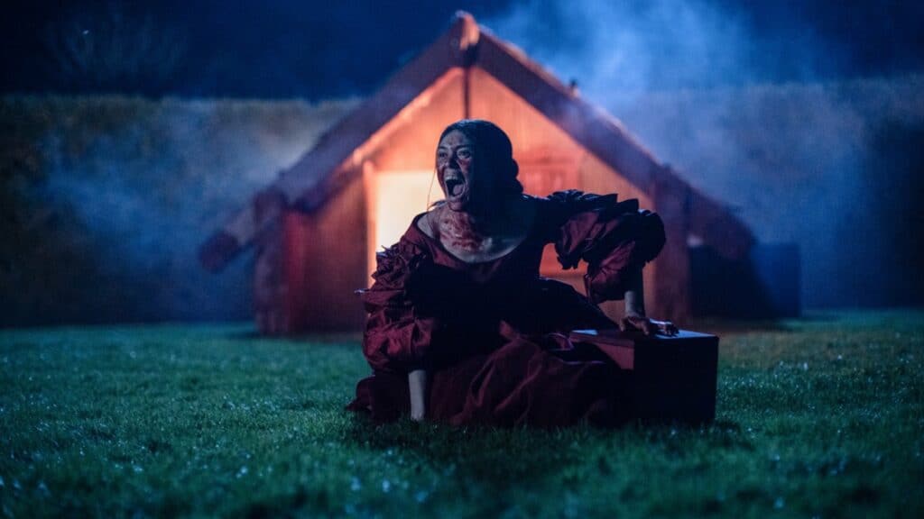 A woman in a bloodstained dress kneels on grass at night, screaming, with a red-lit wooden house in the background.