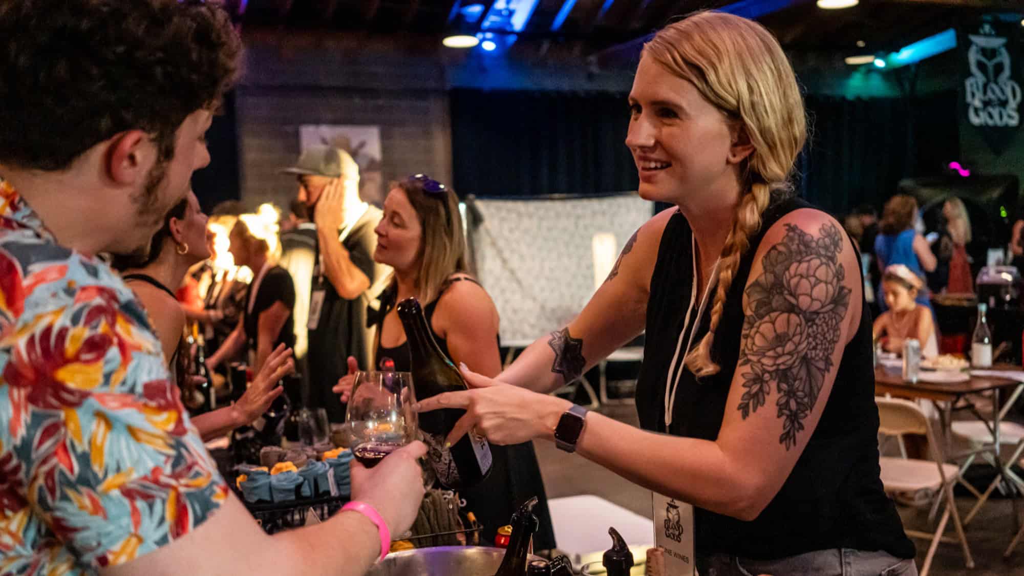 A woman with a floral tattoo serves a drink to a man at a busy indoor event, where the atmosphere buzzes like the vibrant scenes from Grapes of Wrath, with other people and booths lively in the background.