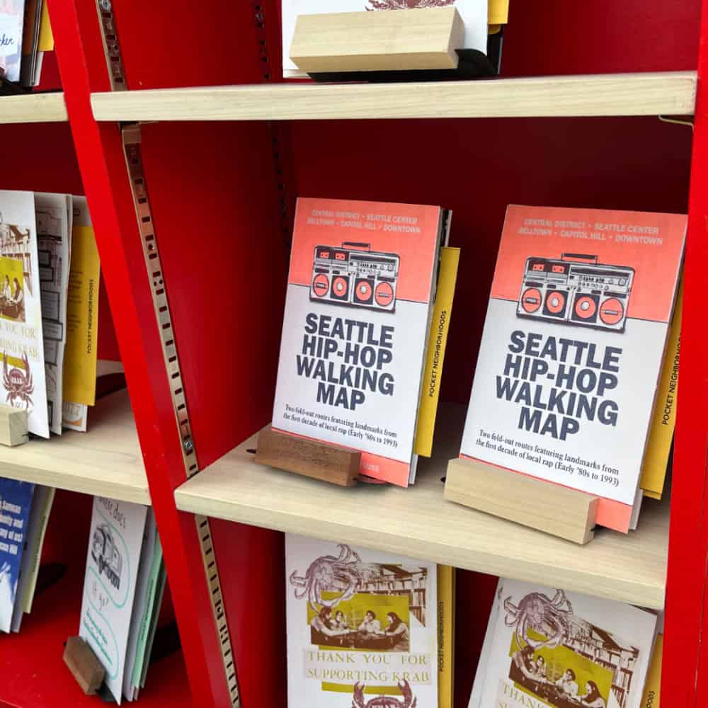 Books titled "Seattle Hip-Hop Walking Map" are displayed on a shelf, alongside pamphlets highlighting Seattle food and culture and things to do in Seattle, including gems like Pike Place Market.