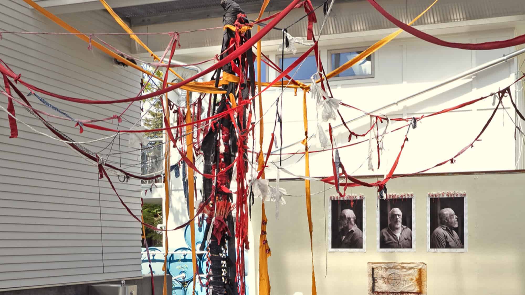 Colorful red, yellow, and orange fabric strips are strung across an outdoor space near a wall displaying three black-and-white portrait photos from Timothy White Eagle's Studio Sessions.