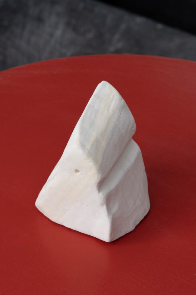 A white, angular stone sculpture with smooth and rough surfaces sits on a red tabletop.