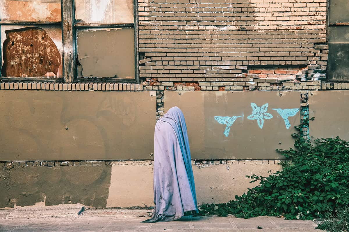 A person draped in a light grey sheet stands against a worn brick wall with broken windows and blue graffiti of two birds and a flower, evoking the gritty charm of Pioneer Square’s urban landscape photography. Green plants grow in the bottom right corner.