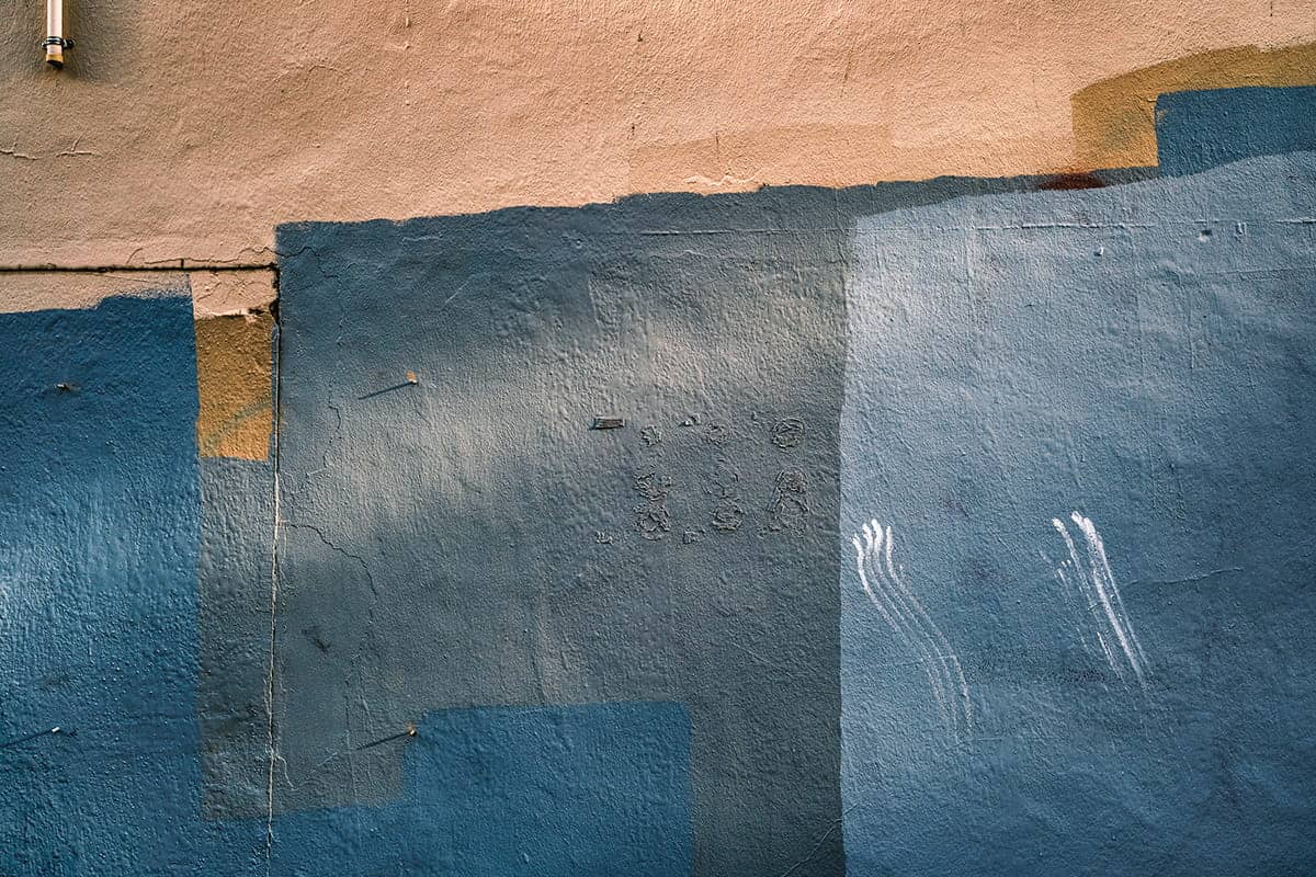 A wall in Pioneer Square with patches of blue, beige, and gray paint, faint markings, and white streaks, showing layers of weathering and repairs—bearing silent witness to Seattle graffiti and the stories of homelessness in Seattle.