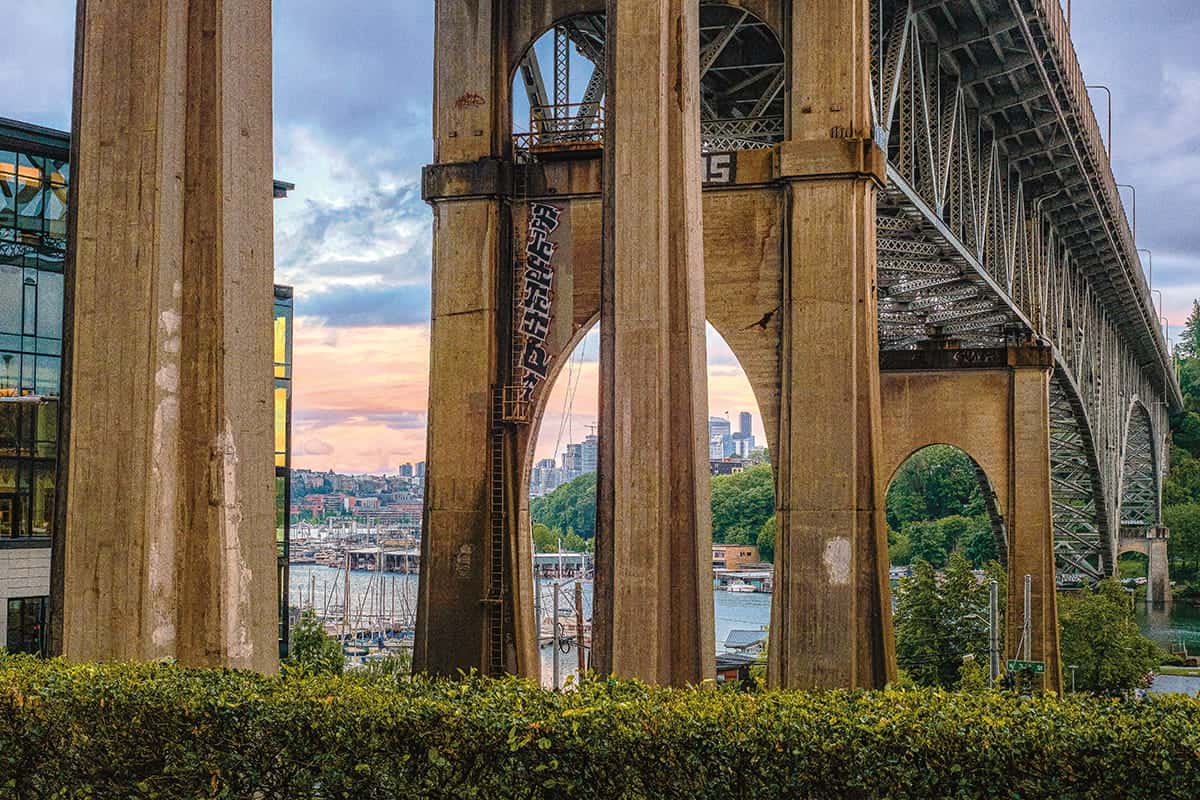 Large bridge with concrete pillars crosses over water at sunset; city buildings, boats, and greenery—plus touches of Seattle graffiti—add urban color to the background.