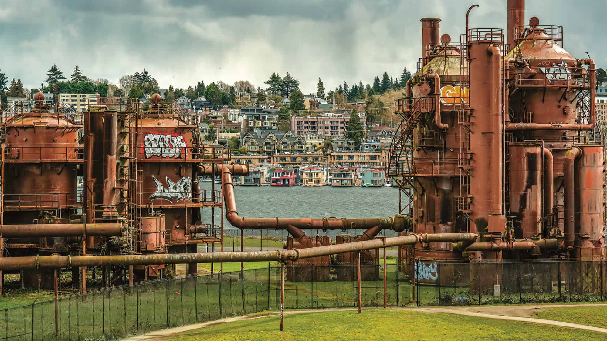 Rusty industrial structures with Seattle graffiti stand on green grass near a body of water, with houses and trees in the background under a cloudy sky.