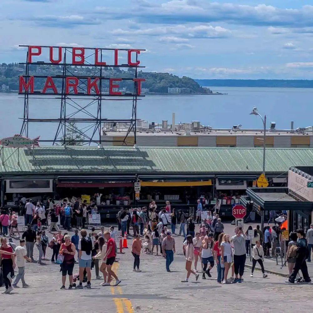 Crowd of people walking in front of the Pike Place Market sign with a view of the water and distant greenery in the background.