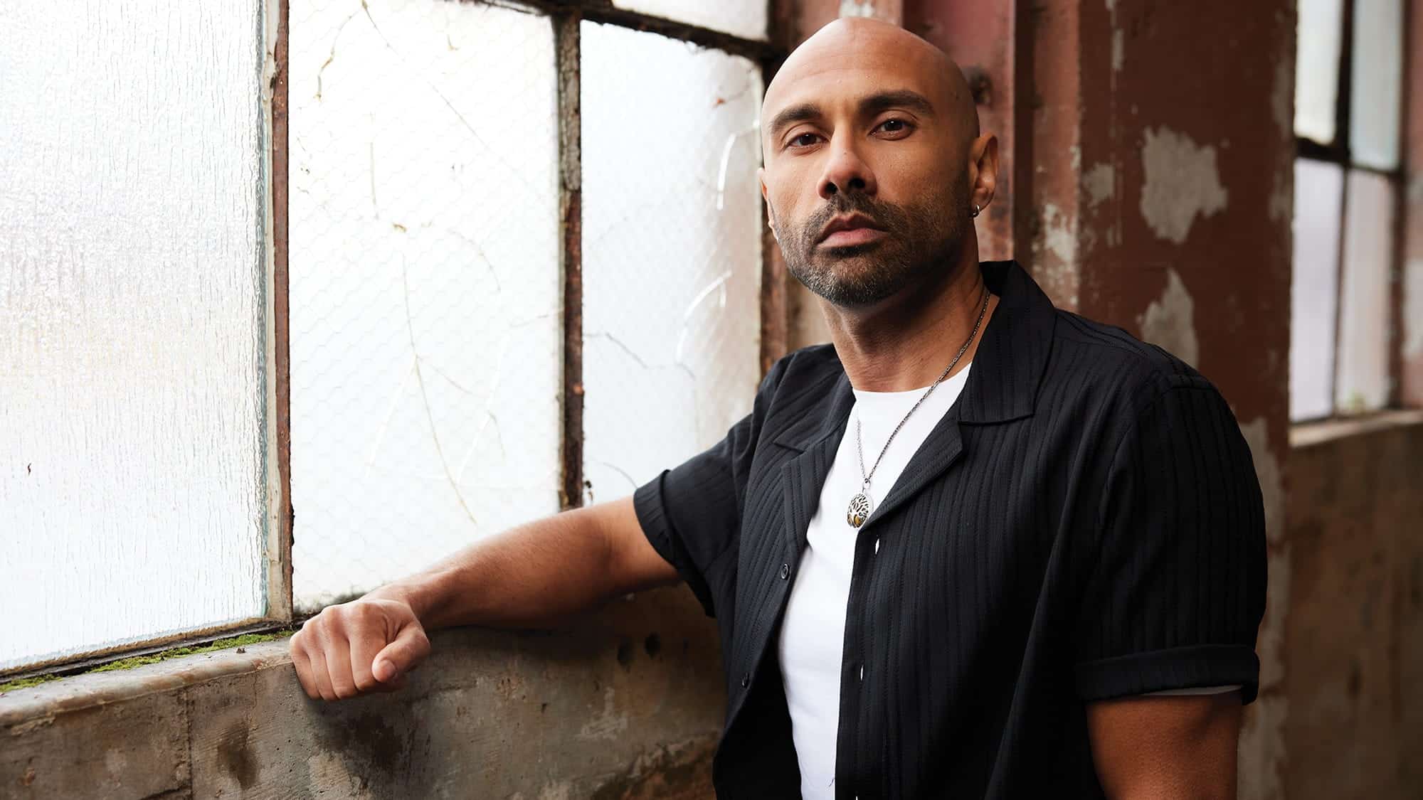 A man with a shaved head and beard stands indoors by a worn window, sporting a black short-sleeve shirt over a white tee and a necklace—someone who could be sharing stories of the justice-impacted community on an Unincarcerated Productions podcast.