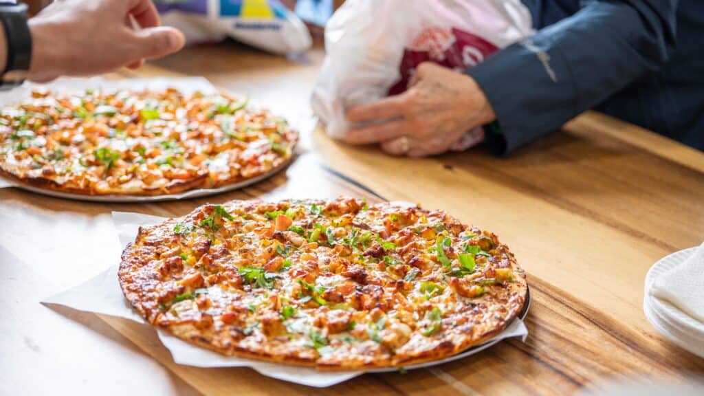Two thin-crust pizzas topped with chicken and herbs sit on a wooden table, as hands reach for food passports and other items in the background.