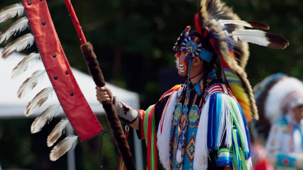 A person in colorful traditional regalia and headdress holds a feathered staff and red flag during an outdoor film festival.