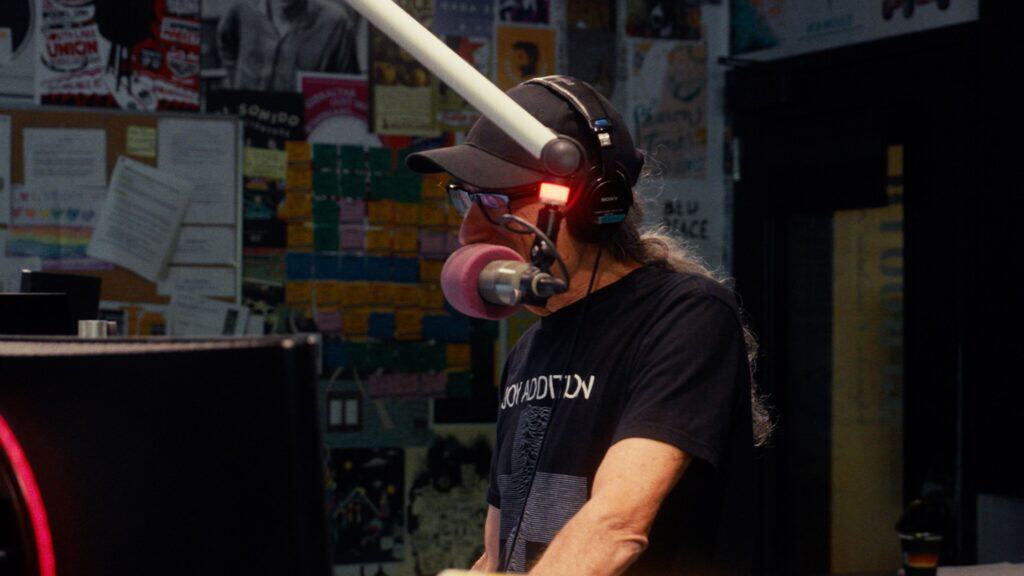 A person wearing headphones and speaking into a microphone stands in a radio studio filled with posters, notes, and memorabilia from SIFF Movies on the walls.