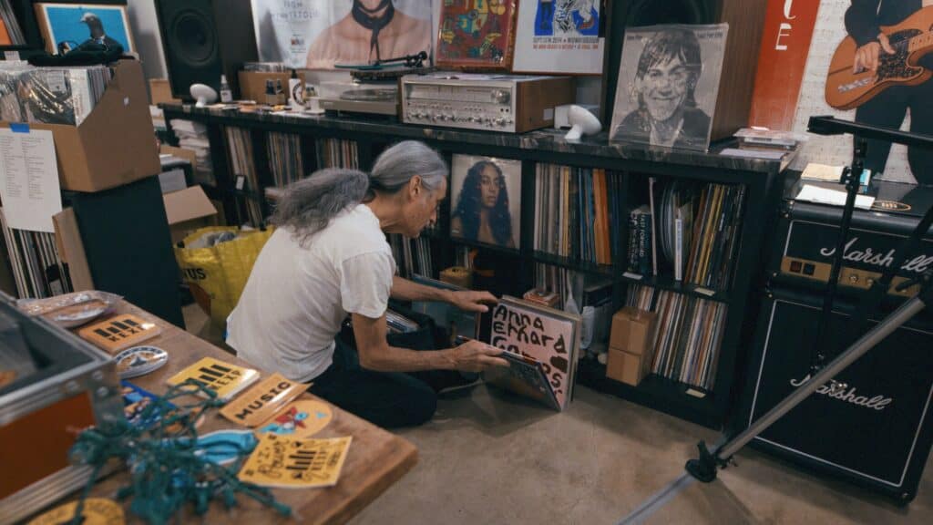 An older person with long gray hair kneels in front of a shelf of vinyl records, browsing through albums in a room filled with music equipment and various items.
