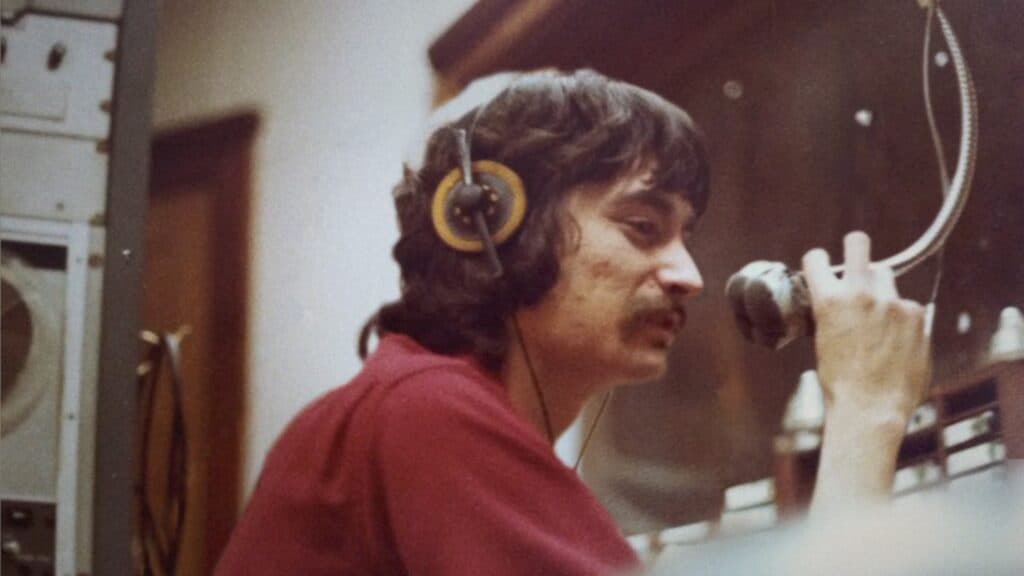 A man with dark hair and a mustache wears headphones and speaks into a microphone in a recording or radio studio.