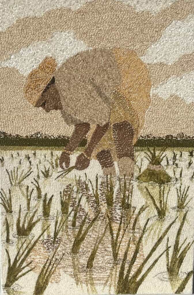 Embroidered artwork of a person bending over to plant rice seedlings in a flooded field under a cloudy sky.