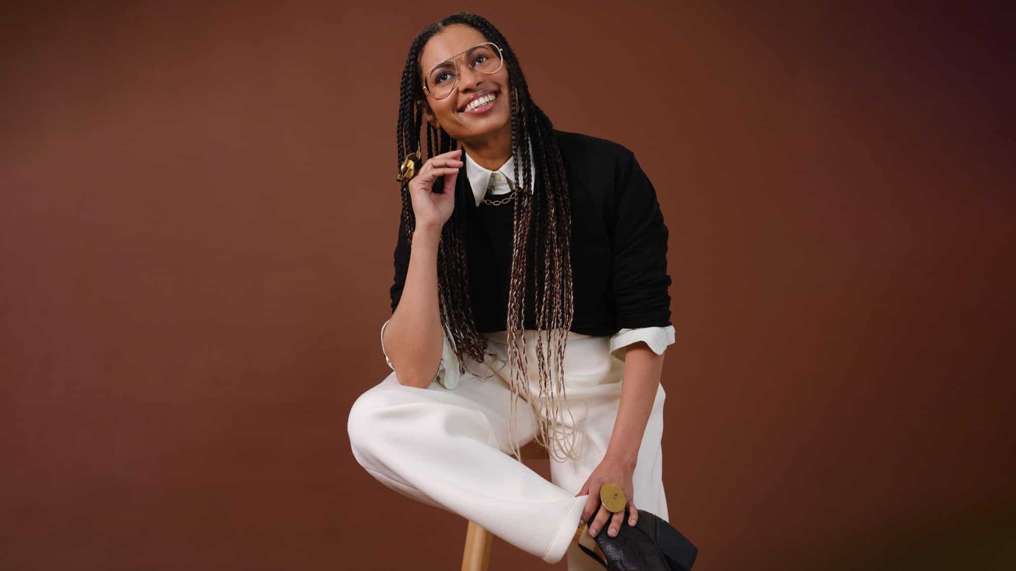 Aina Abiodun, wearing long braids, glasses, and a black sweater over a white collared shirt, sits on a stool against a brown background, smiling and looking upward—an inspiring figure in Seattle climate tech and VertueLab initiatives.