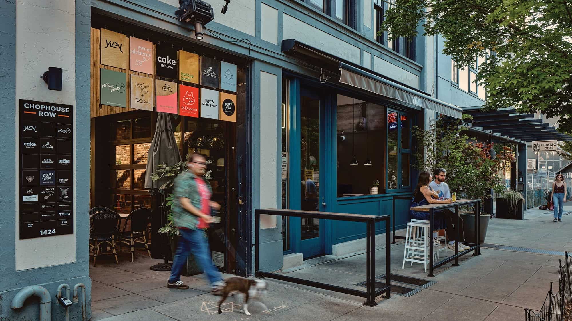 A man walks a dog past a restaurant with open doors along Capitol Hill’s Pike/Pine corridor, while two people sit and talk at a small table outside on the lively city sidewalk.