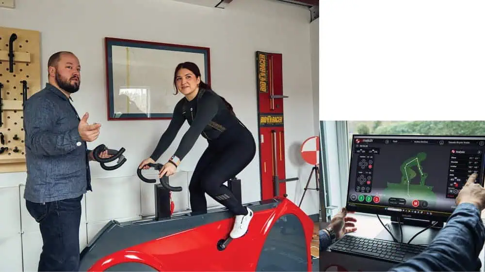 A woman rides a stationary bike indoors at a Fremont bicycle shop while a man speaks to her; a nearby monitor displays an ID Match Bike Lab digital analysis of her cycling posture.