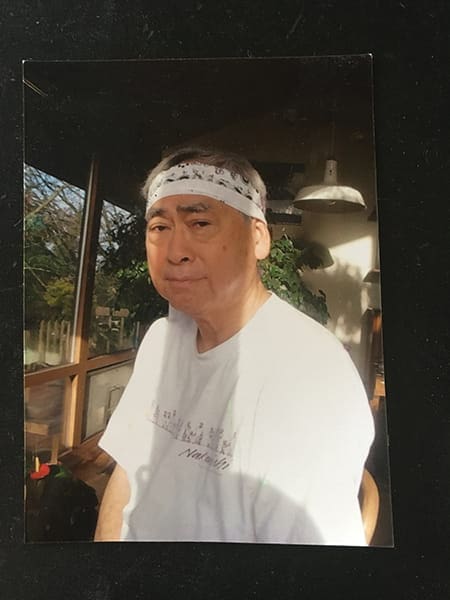 An older man wearing a white T-shirt and a patterned headband sits indoors by a window, sunlight streaming in and plants behind him—a serene moment that could inspire a Seattle Tomodachi comic.