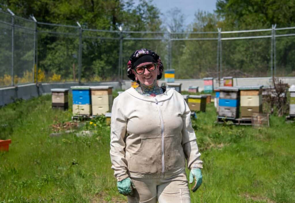 A person wearing a beekeeping suit and gloves stands smiling in front of several colorful beehives in a grassy, fenced area.