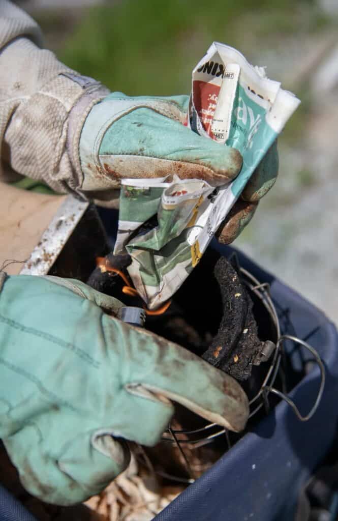 A person wearing gloves crumples a piece of newspaper and places it into a charcoal starter outdoors.