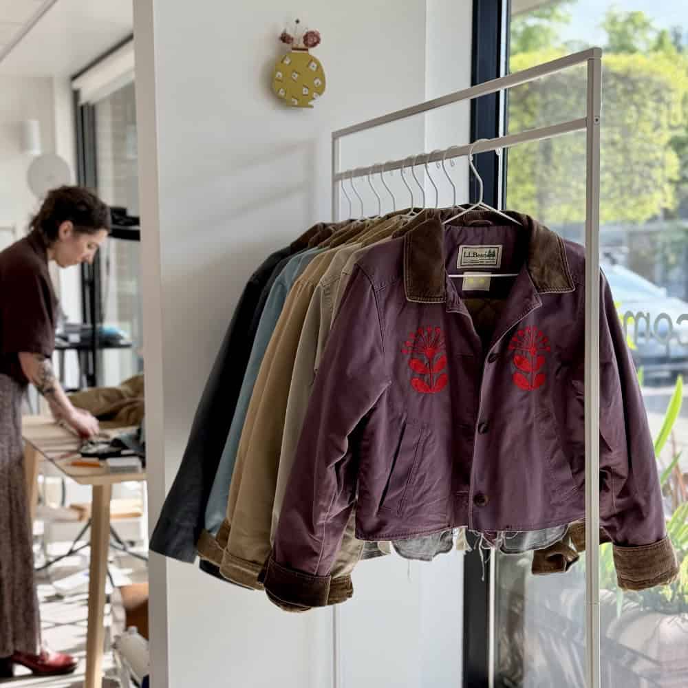 A rack of jackets is displayed in a sunlit room, with a person working at a table in the background near a window.