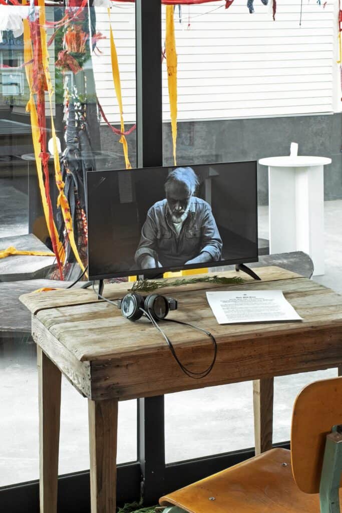 A small wooden table with headphones, a monitor displaying a black-and-white portrait, and a sheet of paper, set by a window with colorful ribbons hanging nearby.