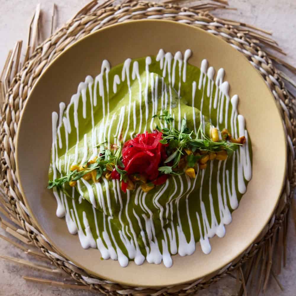 A plate of green sauce-covered enchiladas, garnished with white crema drizzle, pickled red onions, microgreens, and corn, served on a beige plate with a woven placemat.