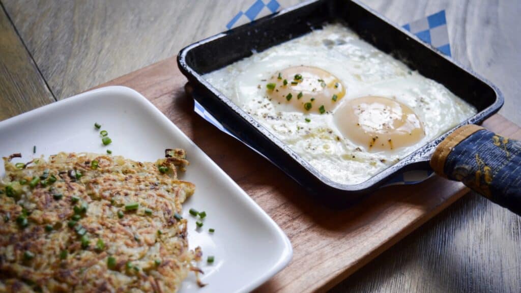 A square skillet with two sunny-side-up eggs garnished with chives next to a plate of browned hash browns on a wooden board.
