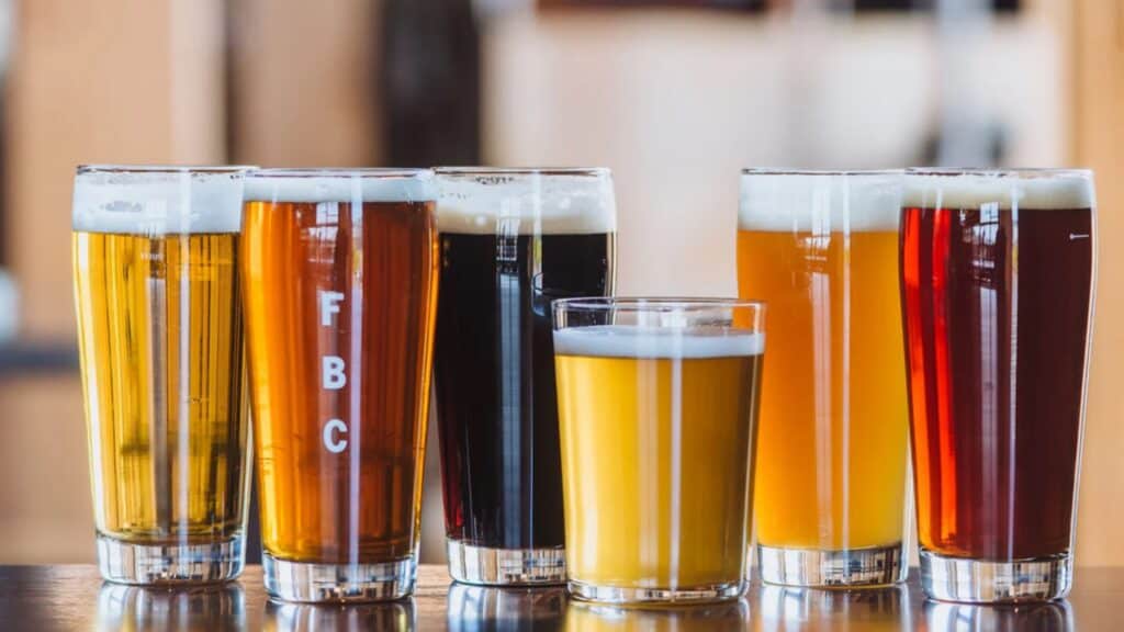 Six glasses of beer in various colors and styles are arranged side by side on a wooden table in Hood River, each with a foamy head—perfect for spontaneous travel with no plan.