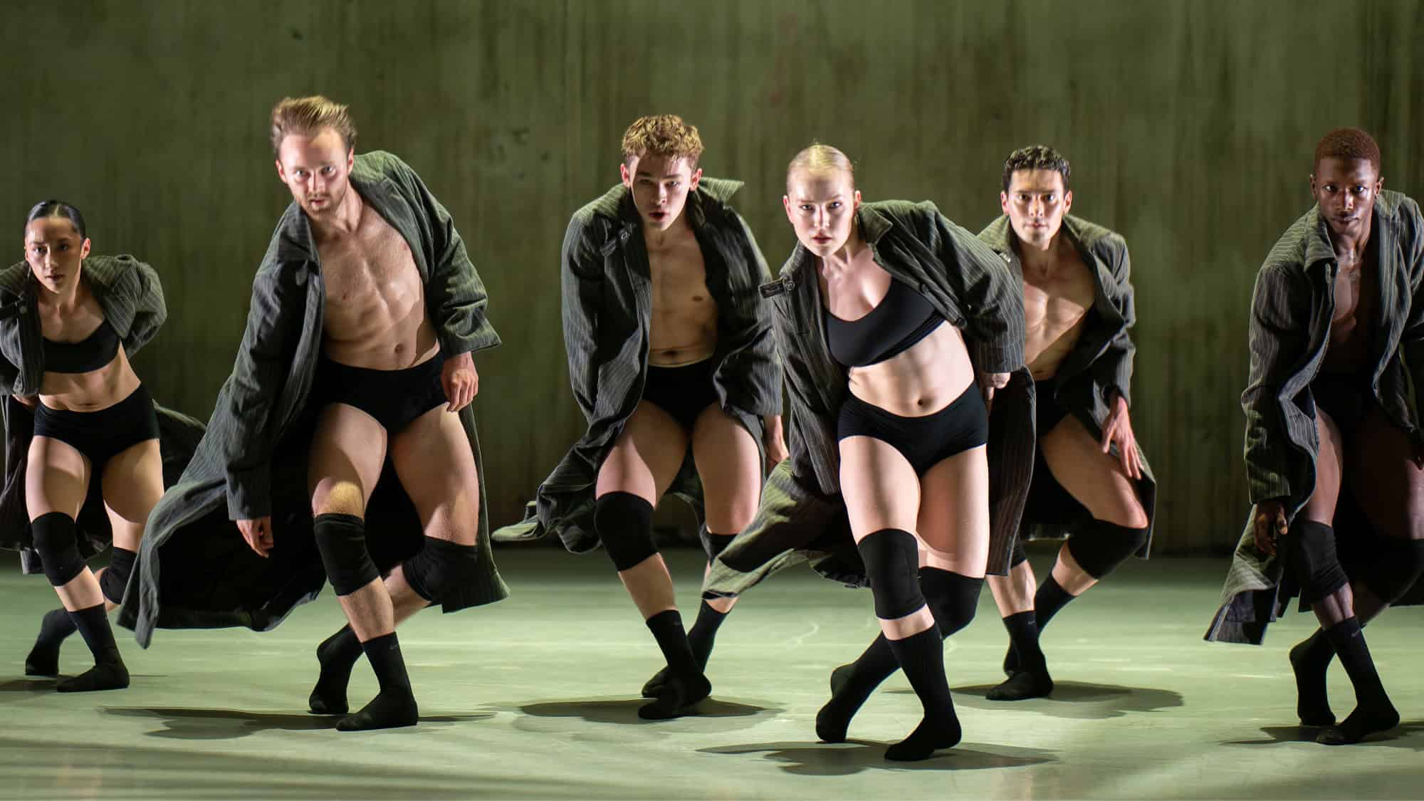 Six dancers in black shorts, tops, and knee pads perform a synchronized routine wearing long open coats on a stage with a plain backdrop.