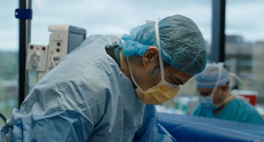 Two surgeons in blue scrubs, masks, hair covers, and gloves perform a procedure in a brightly lit Seattle operating room, with medical equipment visible.