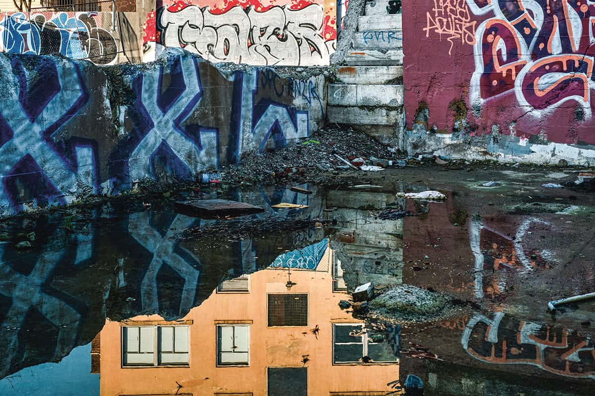 A building’s reflection is visible in a large puddle surrounded by concrete walls and steps covered in colorful graffiti, capturing the raw beauty of urban landscape photography reminiscent of scenes from the More Paint book.