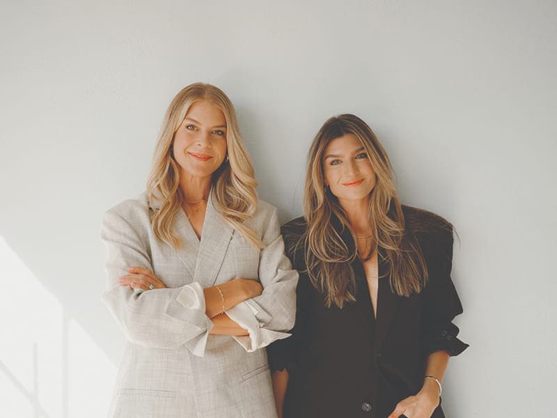Two women stand side by side against a plain light-colored background, both wearing stylish blazers and smiling at the camera, showcasing The Carry vest as an innovative piece of female fitness equipment.