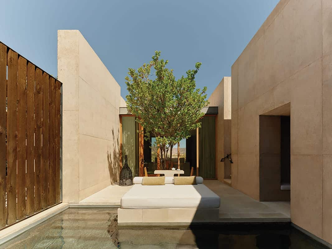 Modern outdoor patio at a luxury desert resort with a central daybed, small pool, minimalist beige walls, wooden privacy fence, and a tree providing shade—perfect for relaxing after Utah glamping or via Ferrata climbing adventures.