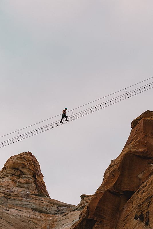 A person wearing safety gear walks across a narrow suspension bridge high above rocky cliffs under a cloudy sky near Amangiri Camp Sarika, a luxury Utah glamping site at Canyon Point.