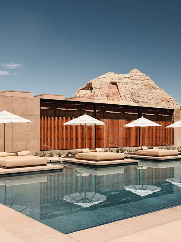 Modern outdoor pool area with cushioned loungers and white umbrellas, set against a contemporary building and rocky desert landscape—perfect for relaxing after a day of via Ferrata climbing at this luxury desert resort.