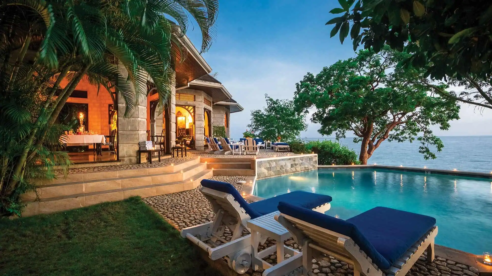 Experience luxury travel Jamaica at Bluefields Bay Villas—a luxury villa with stone exterior, poolside lounge chairs, and an infinity pool overlooking the ocean, surrounded by lush greenery in the evening light.