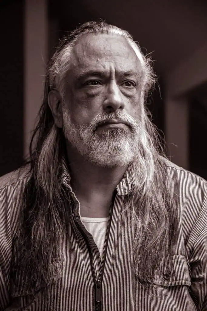 An older man with long gray hair and a beard, wearing a striped shirt and looking thoughtfully into the distance. The image is in sepia tone.