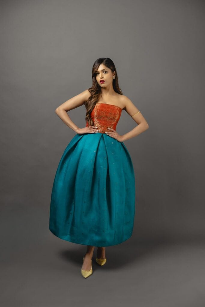 Woman posing in a studio wearing a strapless orange top and a voluminous teal skirt, with yellow high heels, against a plain gray background.