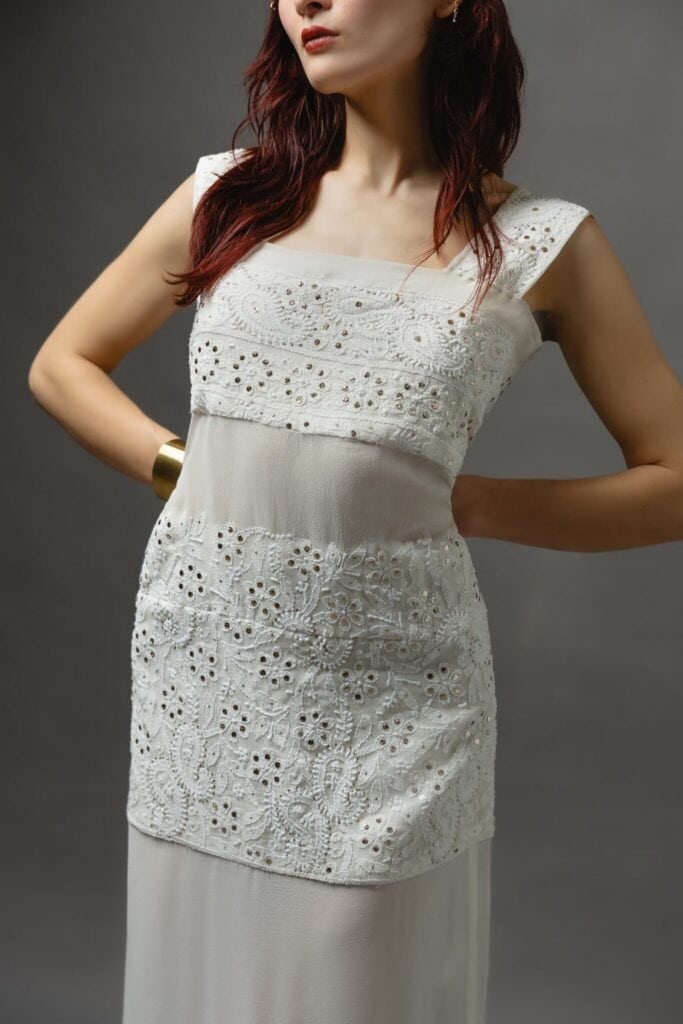 Woman wearing a sleeveless white dress with eyelet lace details, standing against a plain gray background with her hands on her hips.
