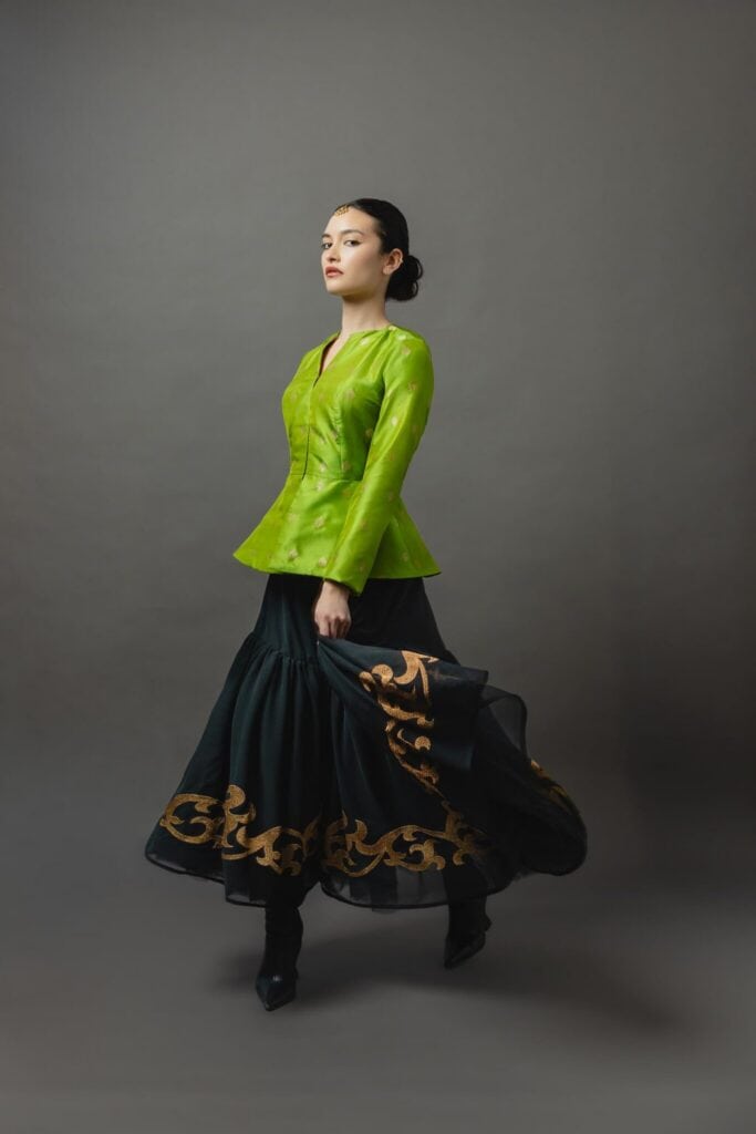 Woman standing against a gray background, wearing a bright green top and a long black skirt with gold embroidery, holding the skirt with one hand as if ready to share the story of her elegant dress.