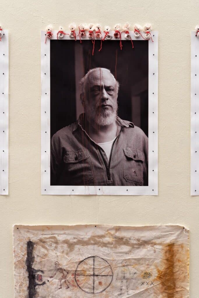 Black and white portrait of an older man with a beard, mounted on a wall with small sculpted heads and red threads above, and a textured drawing displayed below.