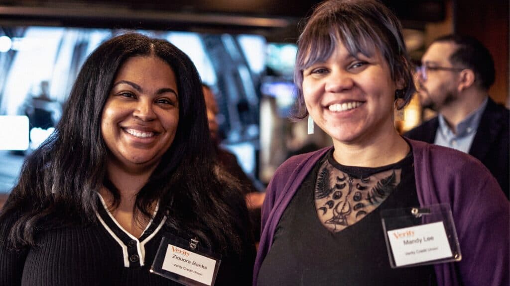 Two women are smiling at an indoor event, each wearing name tags and business attire. Other people are seen in the background.