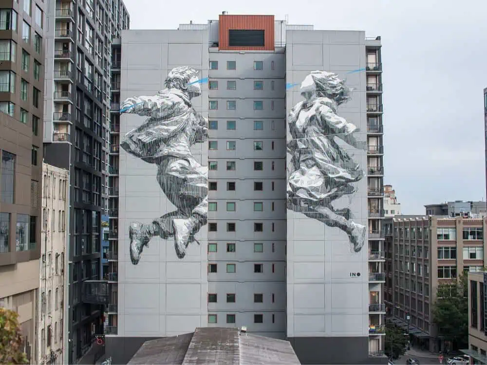 Large mural on a tall building shows two goal-oriented figures, a boy and a girl, leaping towards each other across the structure. Surrounding area features modern apartment buildings.