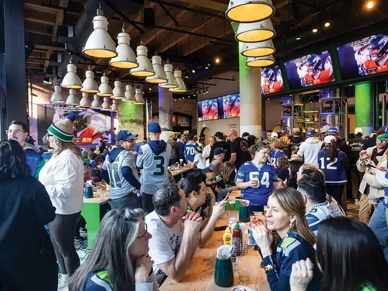 Crowded sports bar in Pioneer Square with people wearing football jerseys, eating and drinking at tables, and watching World Cup games on large TV screens.